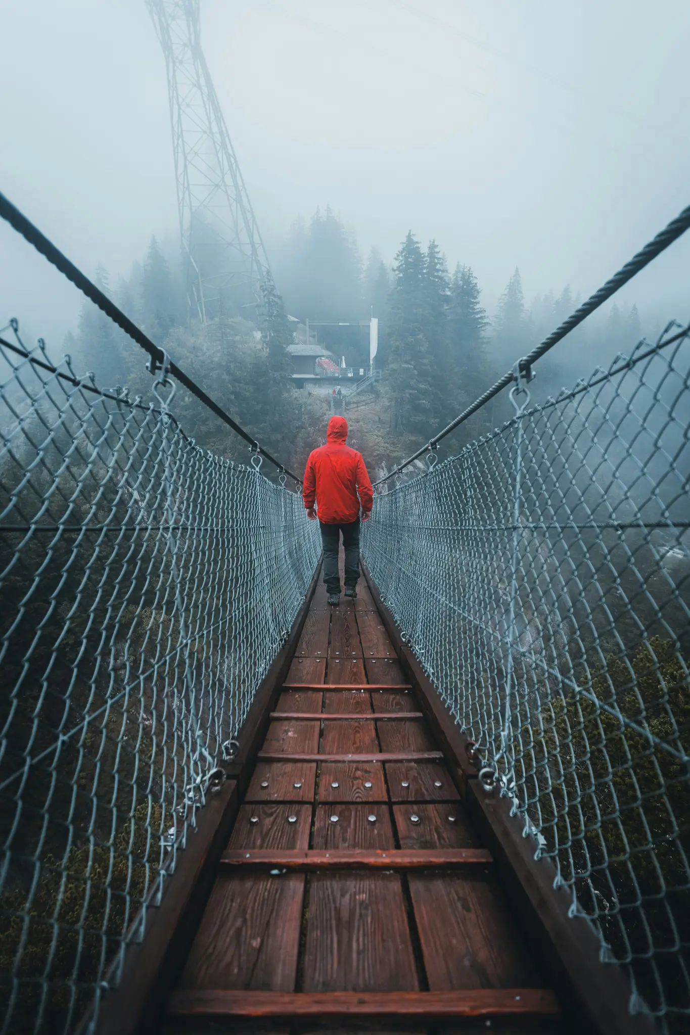 Una persona con una chaqueta roja brillante camina alejándose del espectador por un puente colgante de madera, desapareciendo