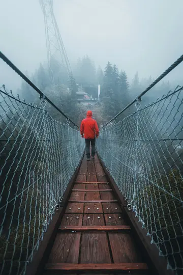 Una persona con una chaqueta roja brillante camina alejándose del espectador por un puente colgante de madera, desapareciendo