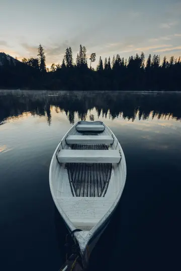 Ein weißes Ruderboot treibt in der Mitte eines ruhigen, dunklen Sees. Im Hintergrund ist ein dichter Wald vor einem