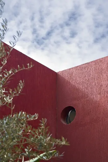 Un edificio rosso con facciata testurizzata e una piccola finestra circolare. Un ulivo con foglie verdi è in primo piano.