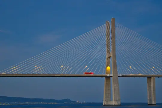 Un puente atirantado con un pilón alto de hormigón gris se alza contra un cielo azul profundo al anochecer. Luces amarillas