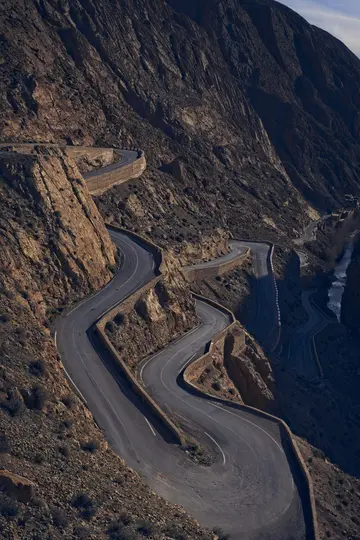 Una vista aérea de una carretera de asfalto oscura con numerosas curvas cerradas, serpenteando por una montaña empinada