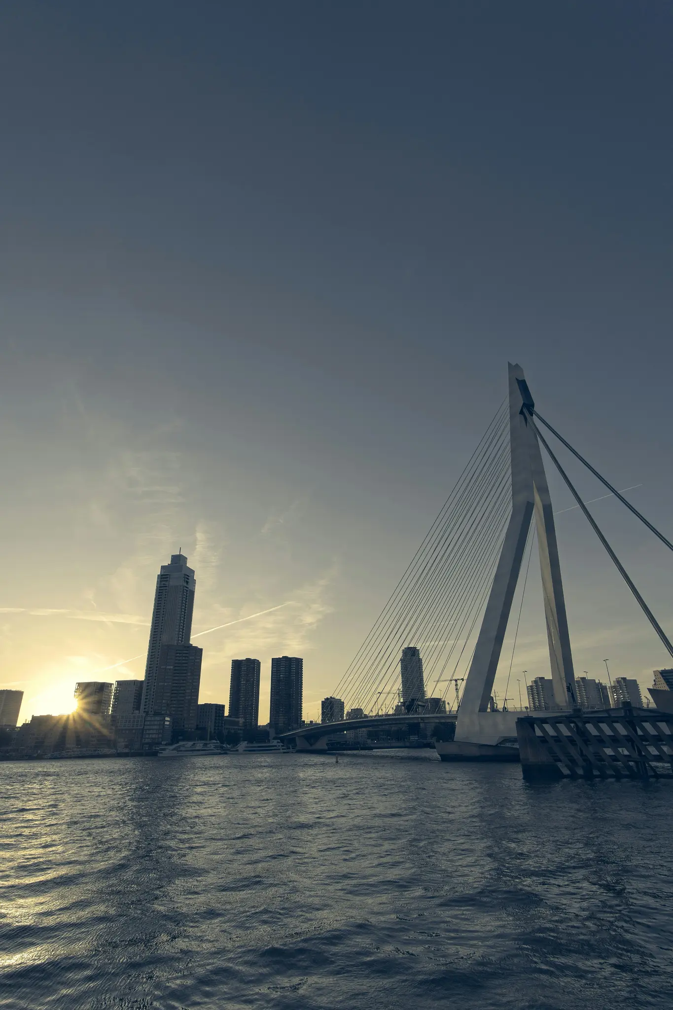 Una toma amplia del skyline de la ciudad y un prominente puente atirantado blanco sobre agua oscura al atardecer. El sol está