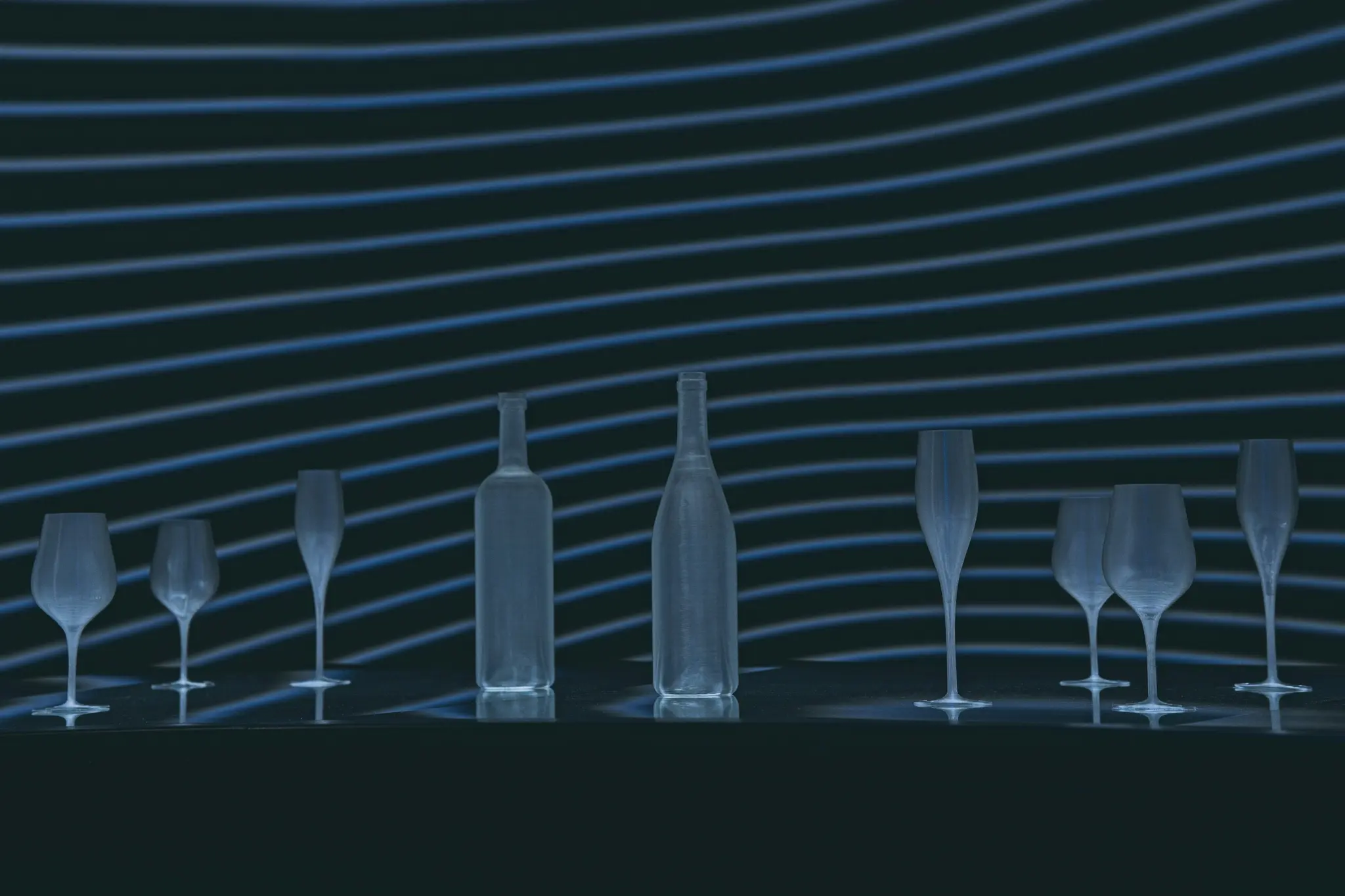 Dos botellas y seis vasos están dispuestos sobre una superficie oscura. El fondo presenta líneas azules horizontales sobre un