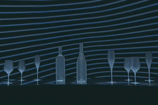 Dos botellas y seis vasos están dispuestos sobre una superficie oscura. El fondo presenta líneas azules horizontales sobre un