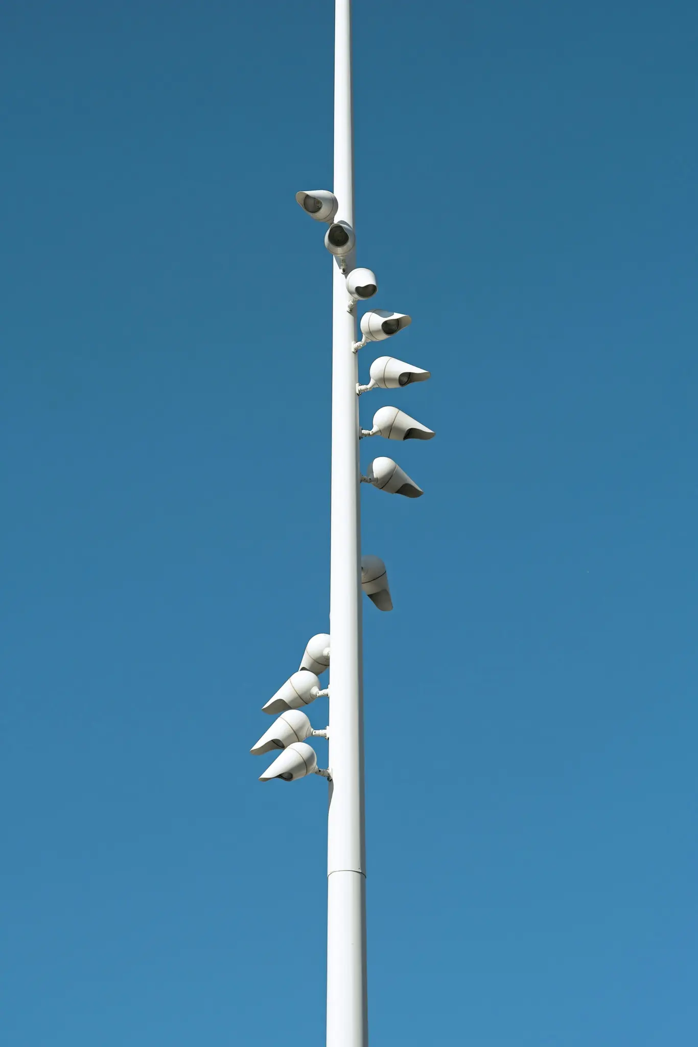 Un poste alto y blanco se erige verticalmente contra un cielo azul claro y brillante. Numerosas cámaras de seguridad blancas