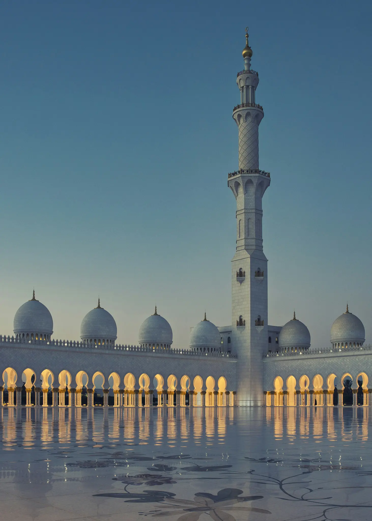 Majesteettinen moskeija hämärässä, jossa on korkea minareetti ja useita kupoleja, ja kultainen valo säteilee kaari-ikkunasta.