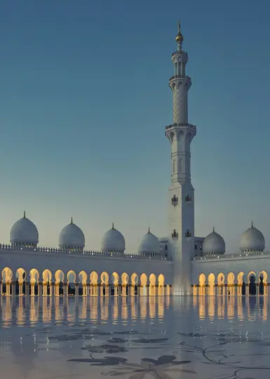 Majestátna mešita za súmraku s vysokou minaretom a niekoľkými kupolami, so zlatým svetlom vychádzajúcim z oblúkového okna.
