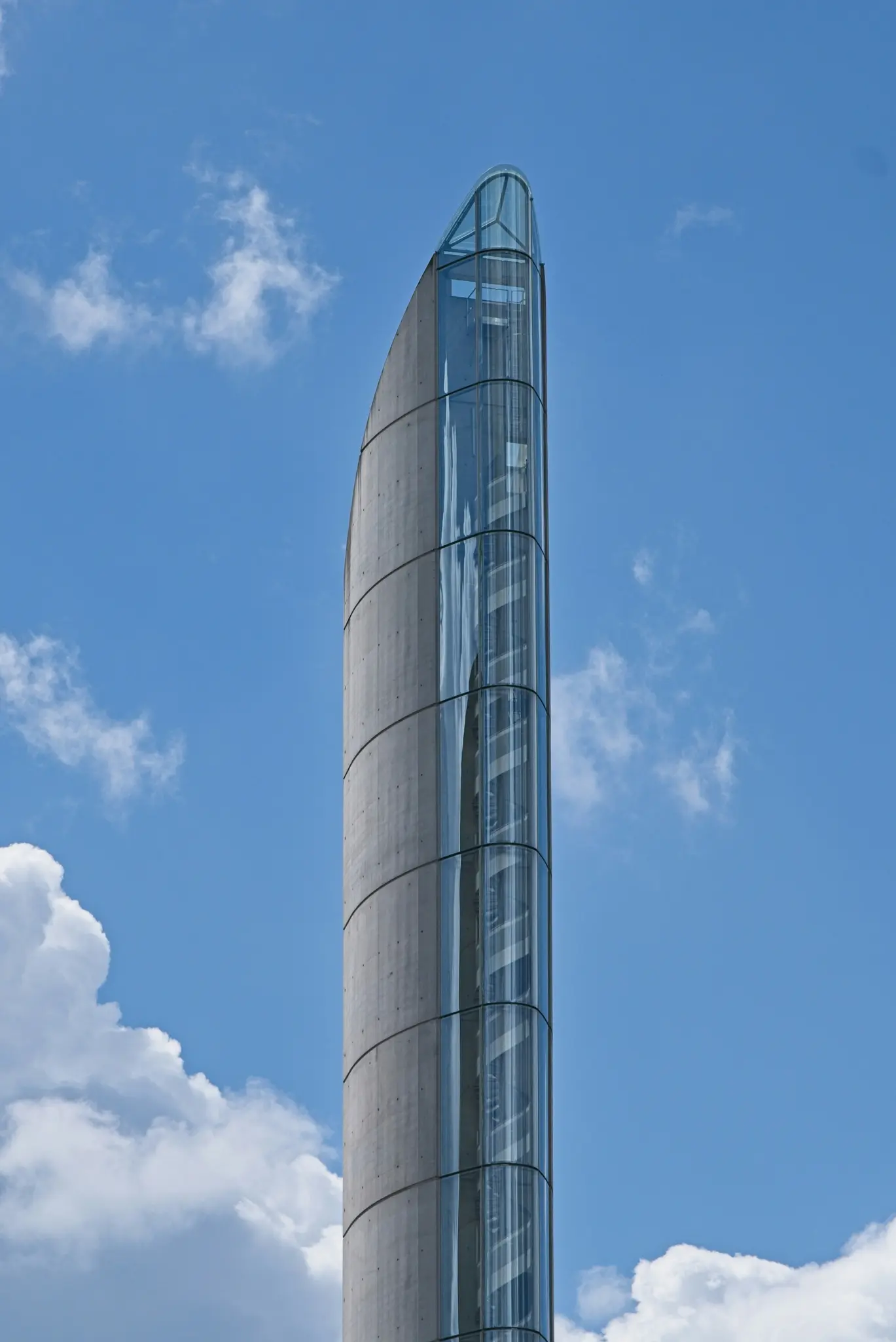 Una torre alta, esbelta y moderna contra un cielo azul con nubes blancas. Un lado es de hormigón curvo, el otro de cristal