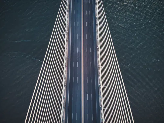Una vista aérea, de arriba abajo, de un moderno puente atirantado. La carretera de asfalto oscura tiene marcas de carril