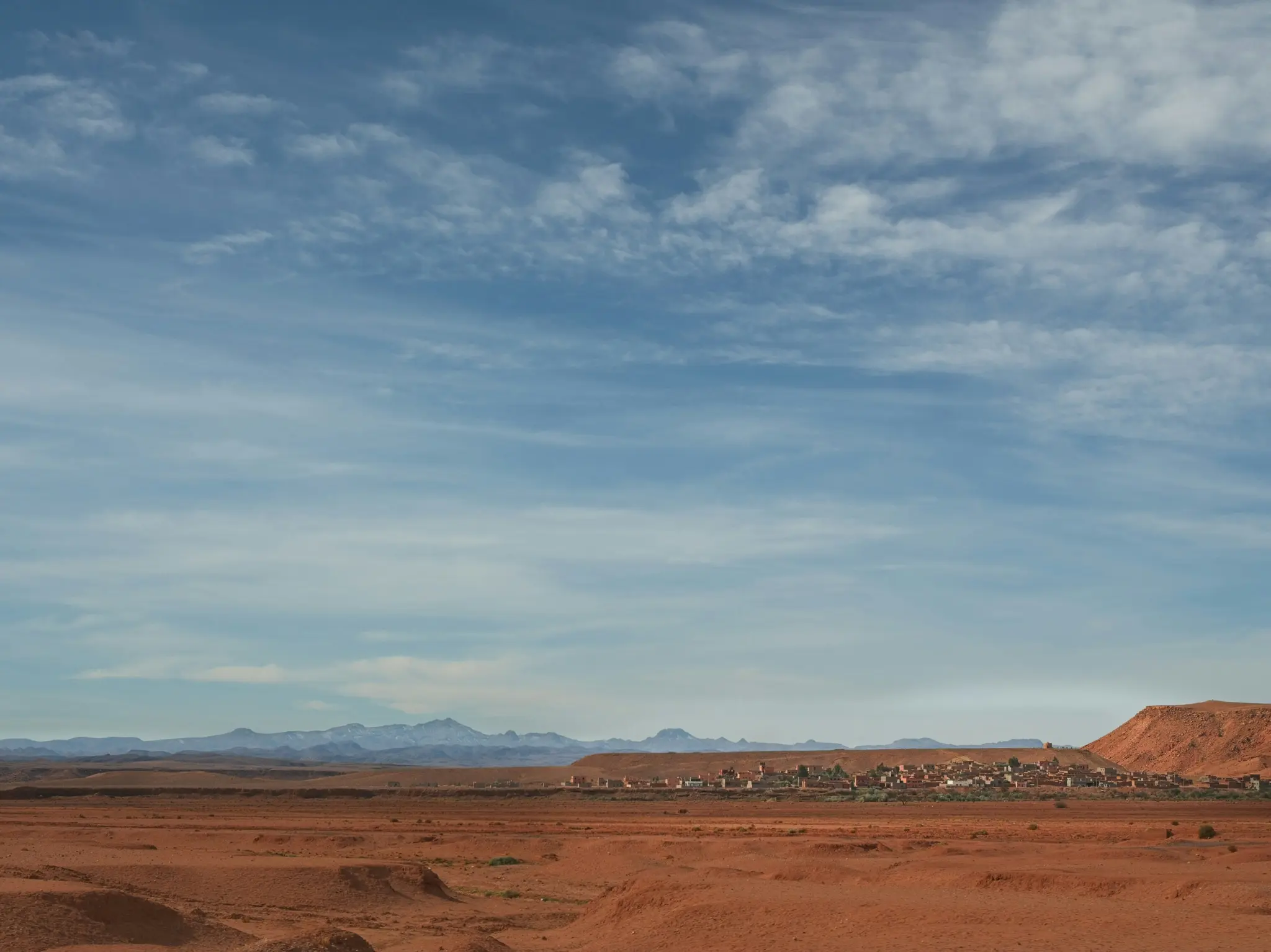 Una toma amplia de un paisaje desértico que presenta un pueblo con edificios de color marrón rojizo, una gran colina roja y