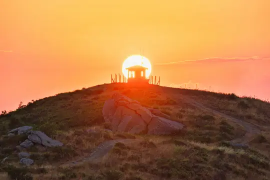 En levende solnedgangsscene med en lille bygning i silhuet på en græsfyldt bakketop. Den store, klare orange sol er direkte…