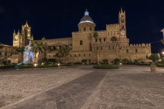 Et vidvinkelbillede af den oplyste Palermo Katedral om natten med dens indviklede arkitektur og kuppel mod en mørk himmel.