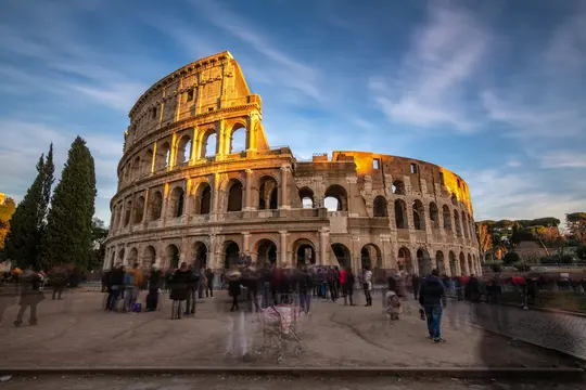 Et vidvinkelbillede af det antikke romerske Colosseum badet i varmt gyldent lys under en blå himmel med fnuggede skyer.