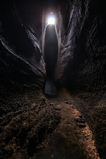 Et lodret billede, der kigger ned ad en meget smal, mørk hulepassage mod et klart, cirkulært lys for enden.