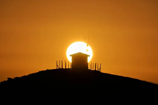 En lille bygning med antenner er silhuet på en bakketop mod en stor, klar orange og gul sol.