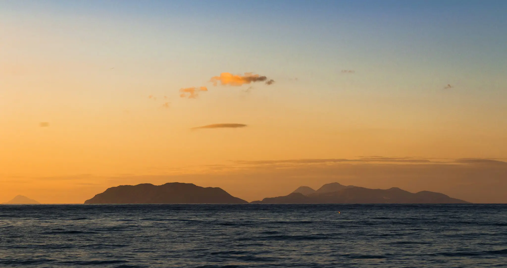 Un'ampia foto panoramica di un tramonto sul mare, con diverse isole scure e in silhouette all'orizzonte. Il cielo è...