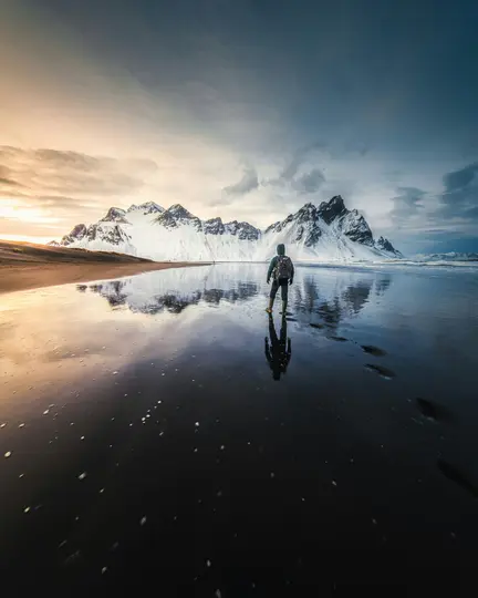 En person står på en våd sort sandstrand og kigger mod snedækkede bjerge under en dramatisk himmel med refleksioner.