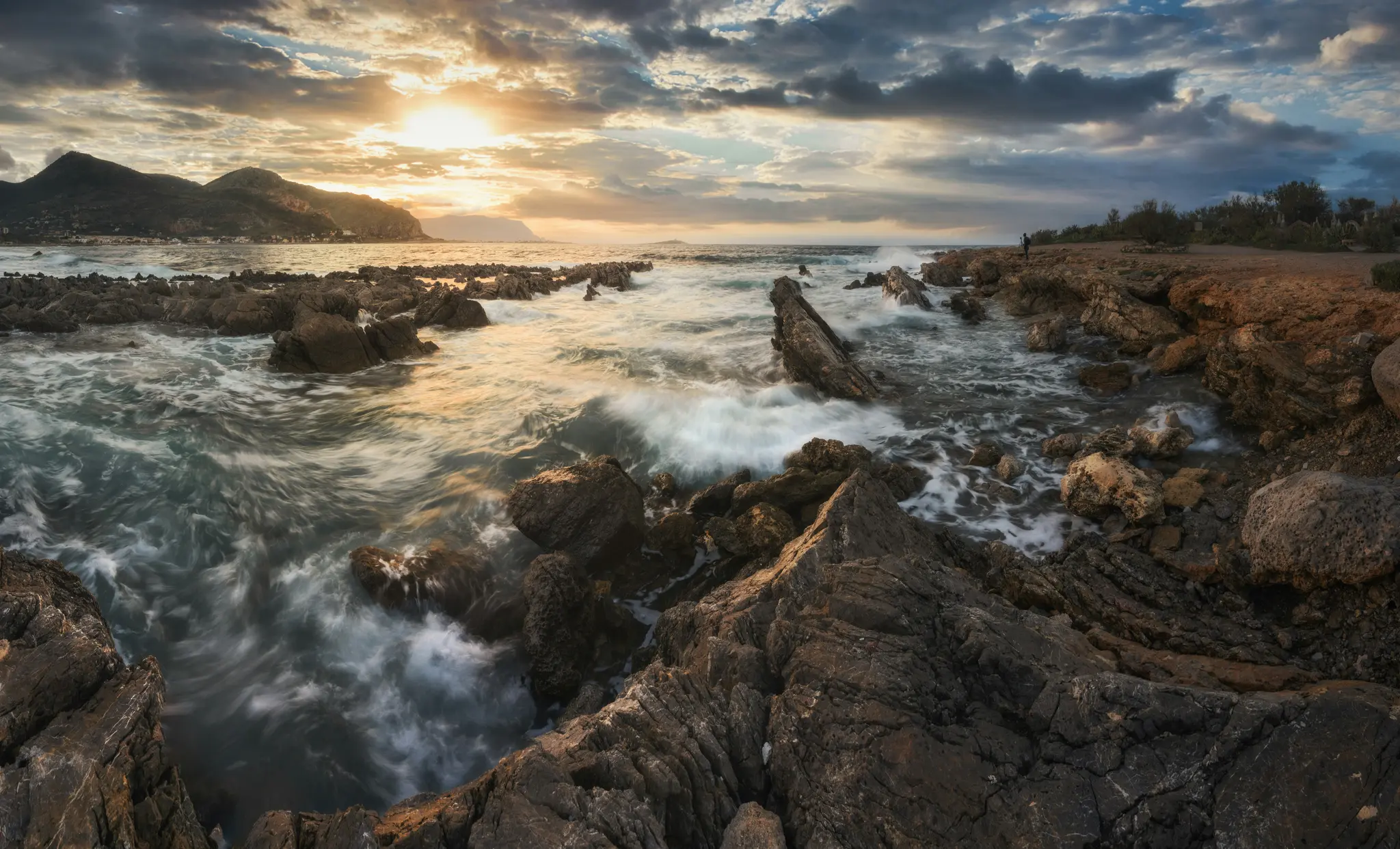 Široká krajinárska fotografia skalnatého pobrežia pri západe slnka. Vlny narážajú na tmavé, zubaté skaly a vytvárajú bielu