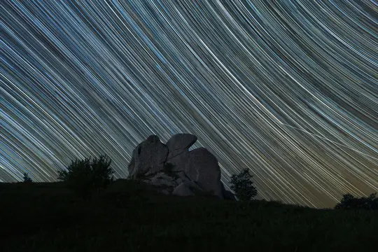 Et langtidsbillede, der viser talrige hvide og gule stjernestier, der buer hen over en mørkeblå nattehimmel. Nedenfor er