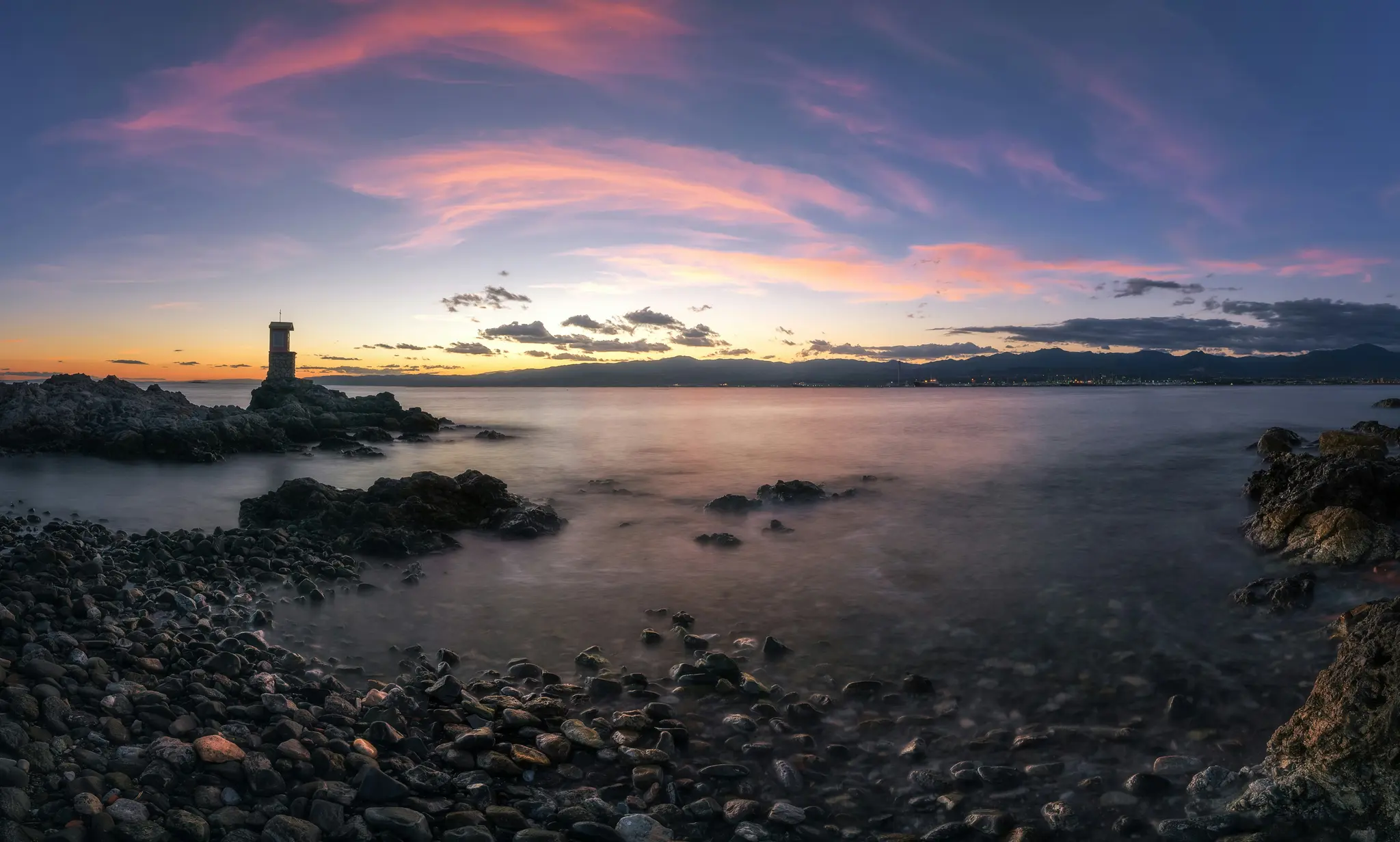 Un'ampia foto panoramica di una costa rocciosa al tramonto. Un piccolo faro scuro si erge su uno sperone roccioso a sinistra.