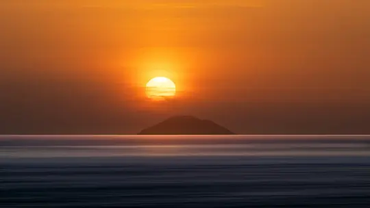 En levende orange og gul solnedgang med solen lavt på himlen, der kaster en lys glød over en mørk, silhuetagtig ø.