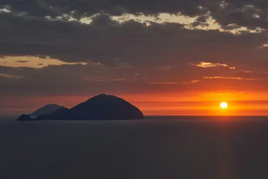 En dramatisk solnedgangsscene med en klar, orange sol lavt på horisonten, der kaster en varm glød over havet. Mørke