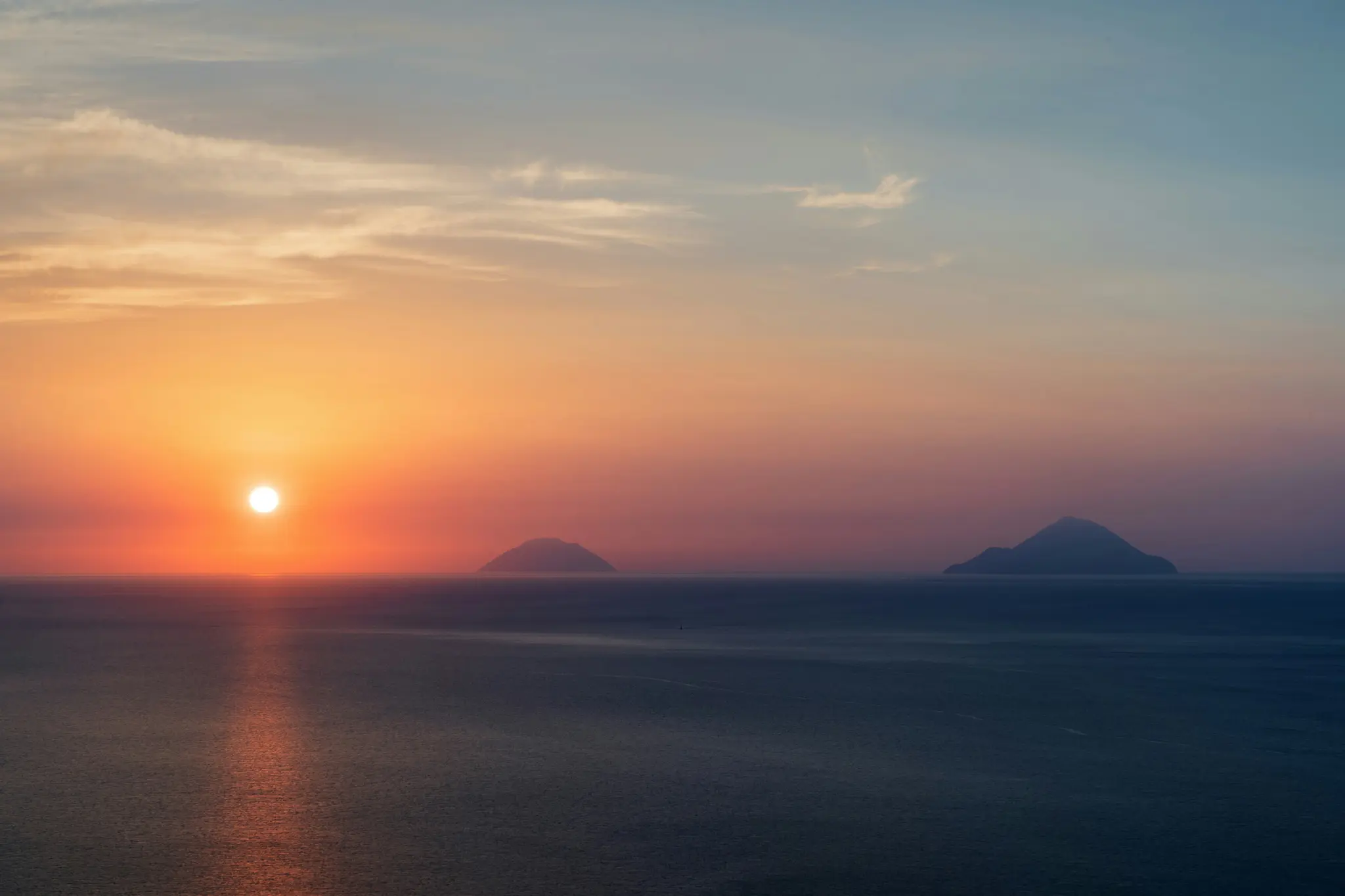 Ett brett landskapsfoto av en levande solnedgång över havet. Solen, en ljus orange sfär, är låg vid horisonten och kastar...