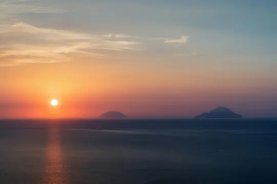 Et bredt landskabsfoto af en levende solnedgang over havet. Solen, en lys orange kugle, er lavt på horisonten og kaster...