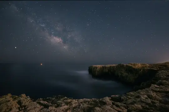 Et langtidsbillede af Mælkevejen, der strækker sig hen over en mørk nattehimmel over en barsk, klippefyldt kystlinje og et