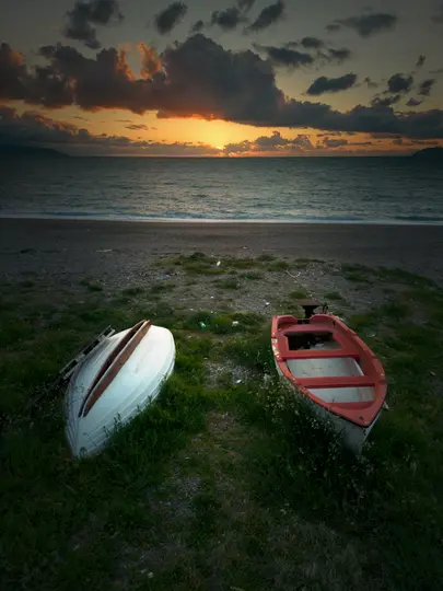 To små både, den ene hvid og væltet, den anden rød og oprejst, hviler på en græsbevokset strand ved solnedgang. Havet er