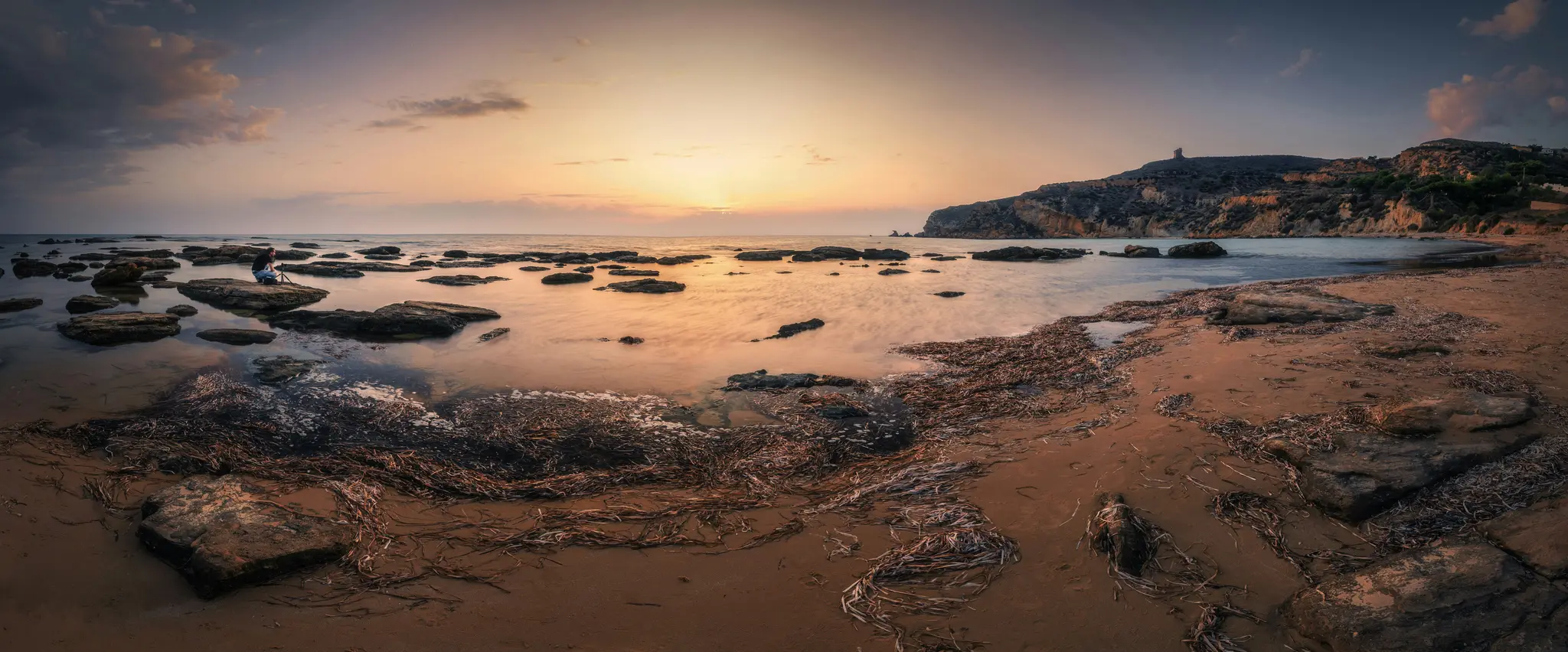Panoramatická fotografia skalnatej pláže pri západe slnka. Obloha je oranžová a modrá. Osoba sedí na kameni v pokojnej vode.