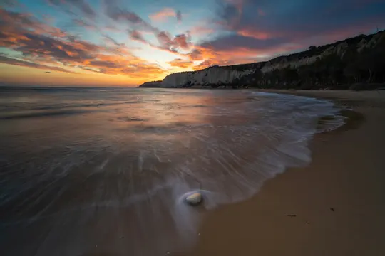 Et langtidsudløserbillede af en strand ved solnedgang. Orange og blå skyer fylder himlen over en mørk klippekant. Bølger