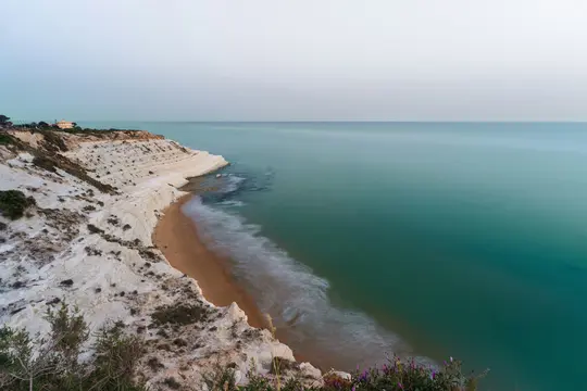 Et langtidseksponeret fotografi af en hvid, lagdelt klippevæg, der bugter sig langs en sandstrand ved siden af et roligt