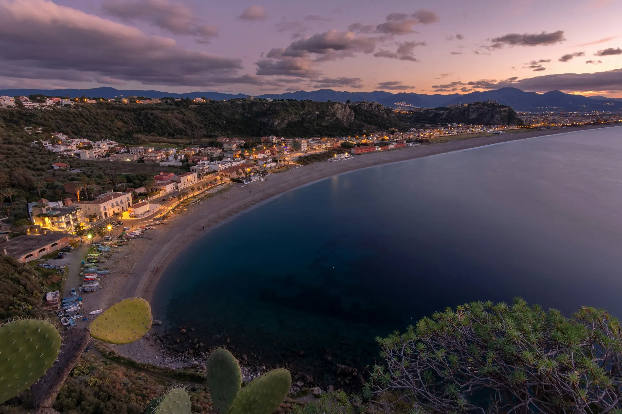 Vista aerea di una cittadina costiera al tramonto, con luci che illuminano gli edifici lungo una spiaggia curva accanto a un