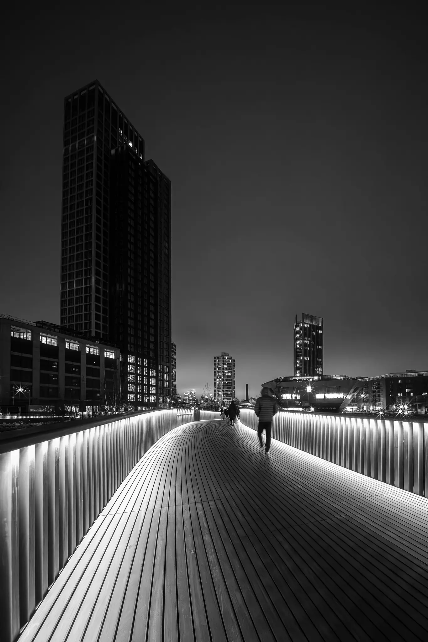 Černobílá fotografie osoby kráčející po jasně osvětleném, zakřiveném dřevěném mostě v noci, obklopené vysokými městskými