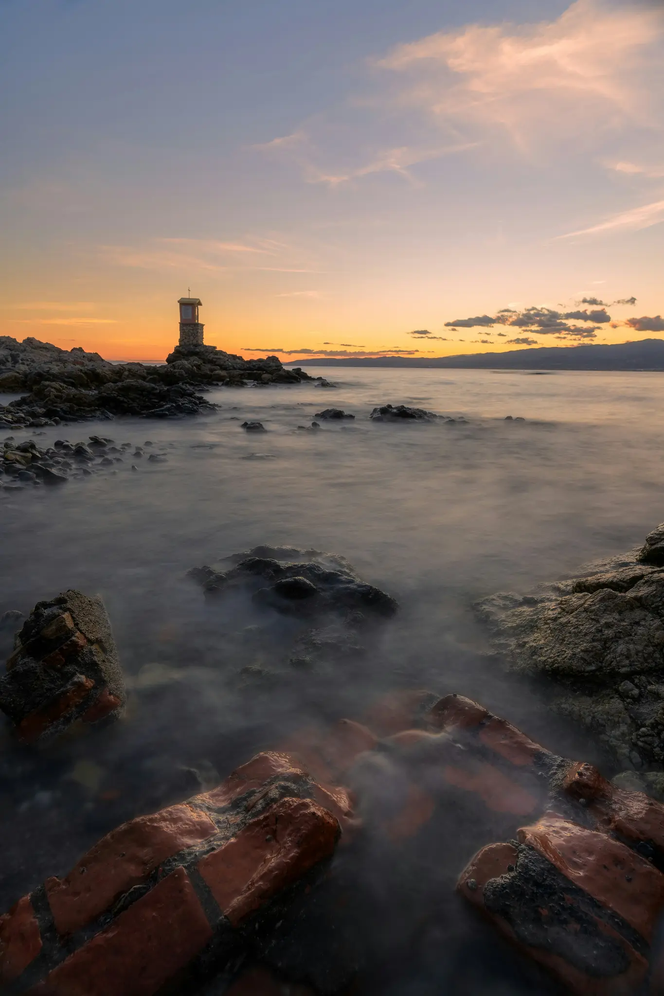 Una fotografia a lunga esposizione di una costa rocciosa al tramonto. Una piccola struttura in pietra con una croce in cima