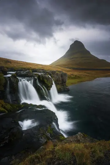 Et langtidsbillede af Kirkjufellsfoss-vandfaldet, der flyder ud i en mørk flod, med det koniske Kirkjufell-bjerg i
