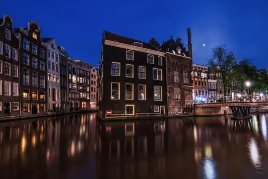Et langtidsbillede af en bykanal om natten, der viser historiske bygninger med oplyste vinduer, der reflekteres i vandet.