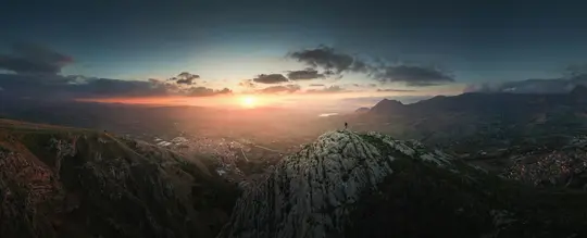 Et panoramabillede af en solnedgang over et stort bjerglandskab. En ensom figur står på en klippetop i midten.