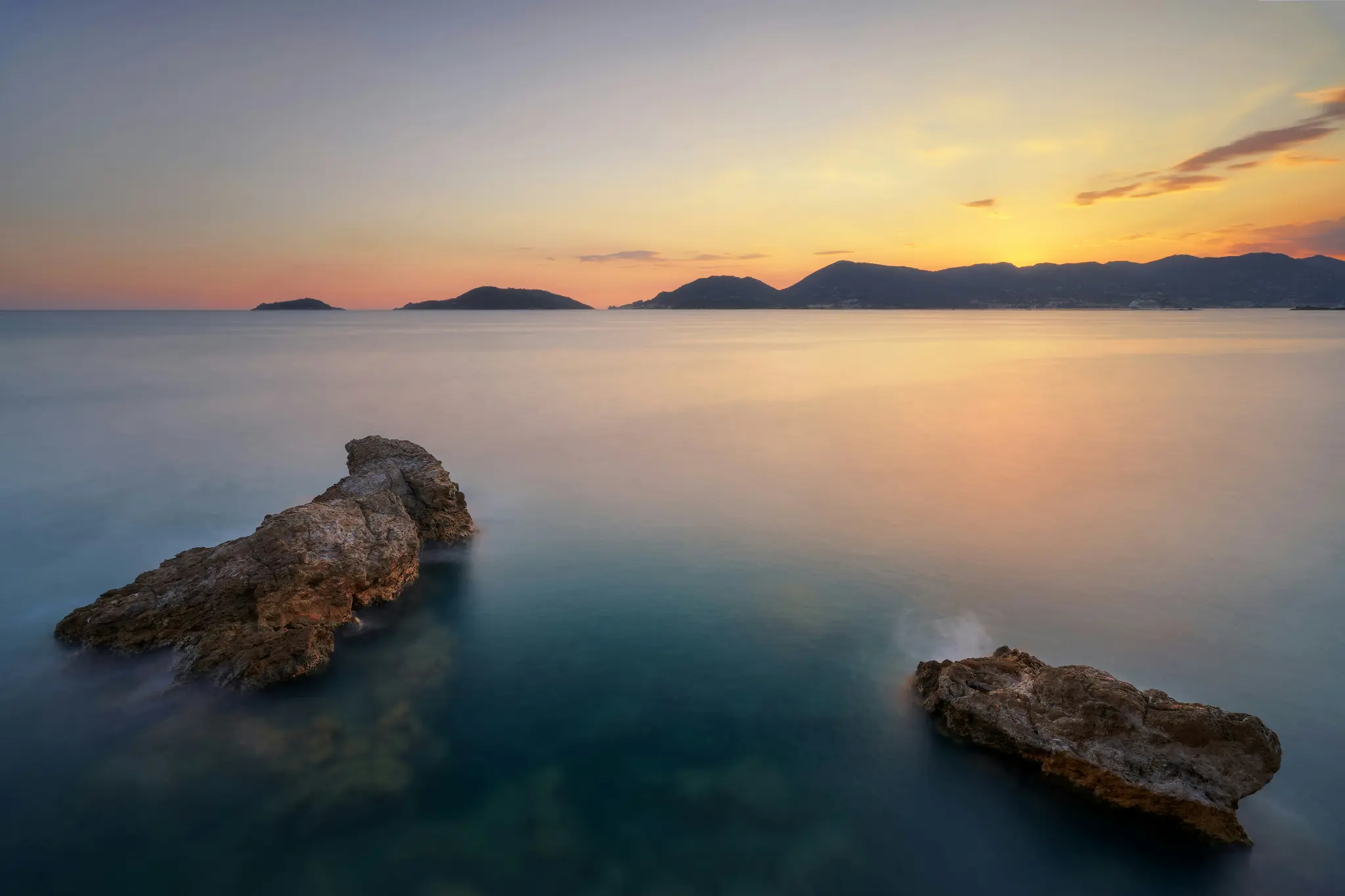 Una foto a lunga esposizione di un tramonto su un mare calmo. Due rocce marroni in primo piano. Isole e montagne lontane