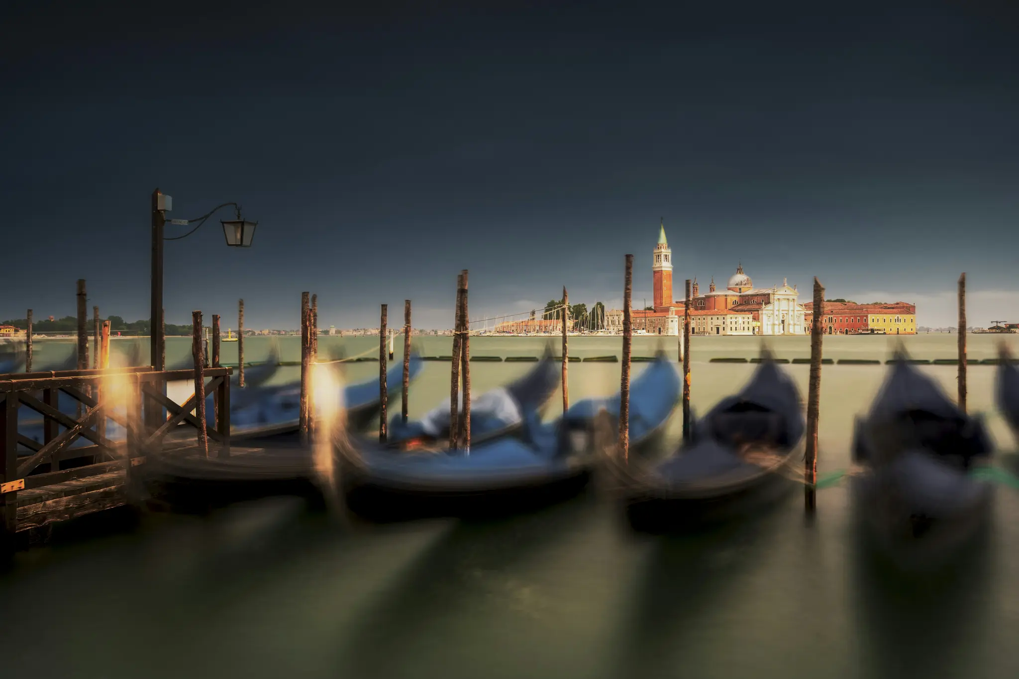 Et langtidseksponeringsfotografi af flere gondoler fortøjet i en venetiansk kanal, med kirken San Giorgio Maggiore og dens
