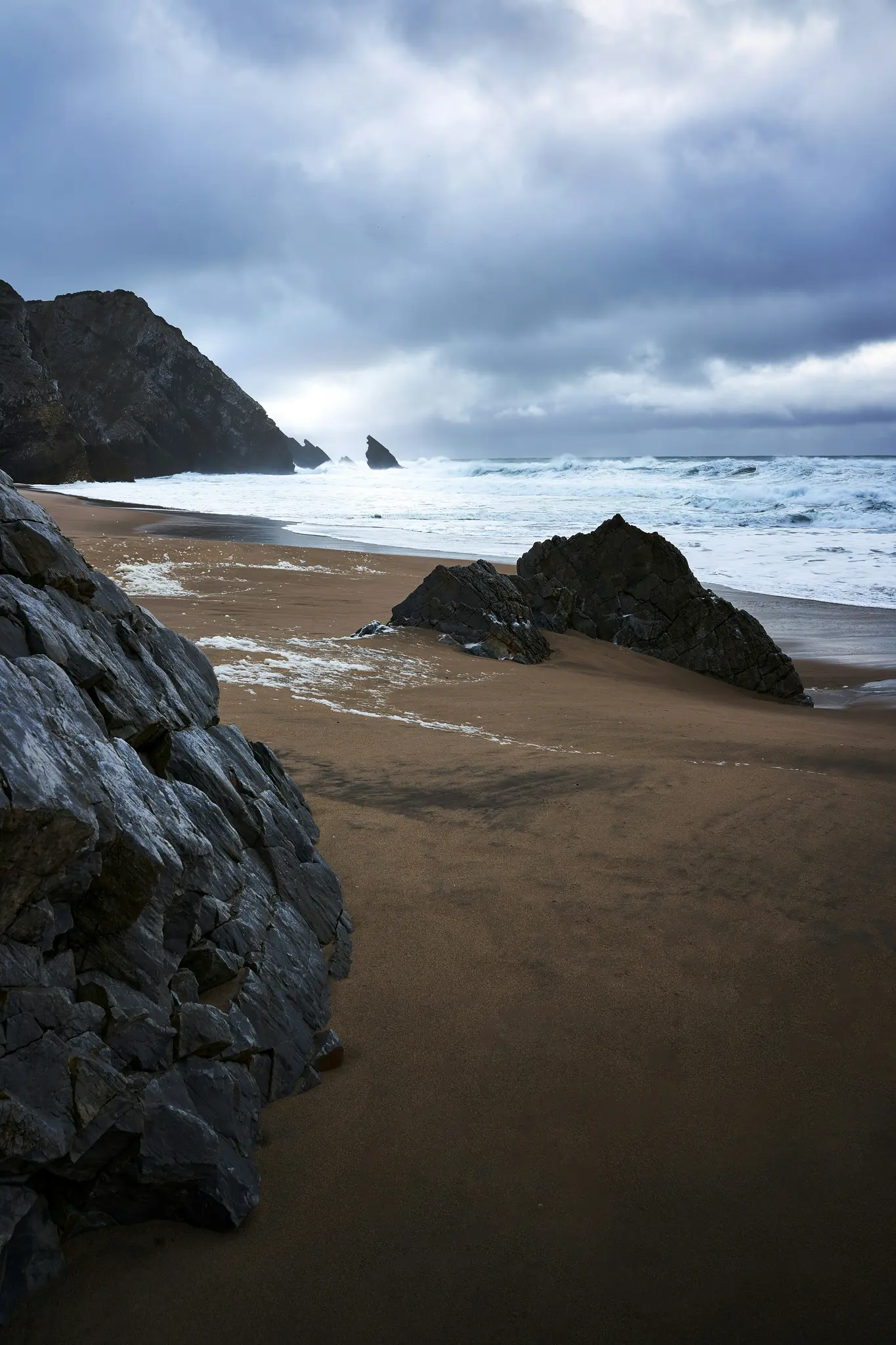 Una ripresa ampia di una spiaggia sabbiosa scura con grandi rocce grigie in primo piano e lungo il lato sinistro. Onde