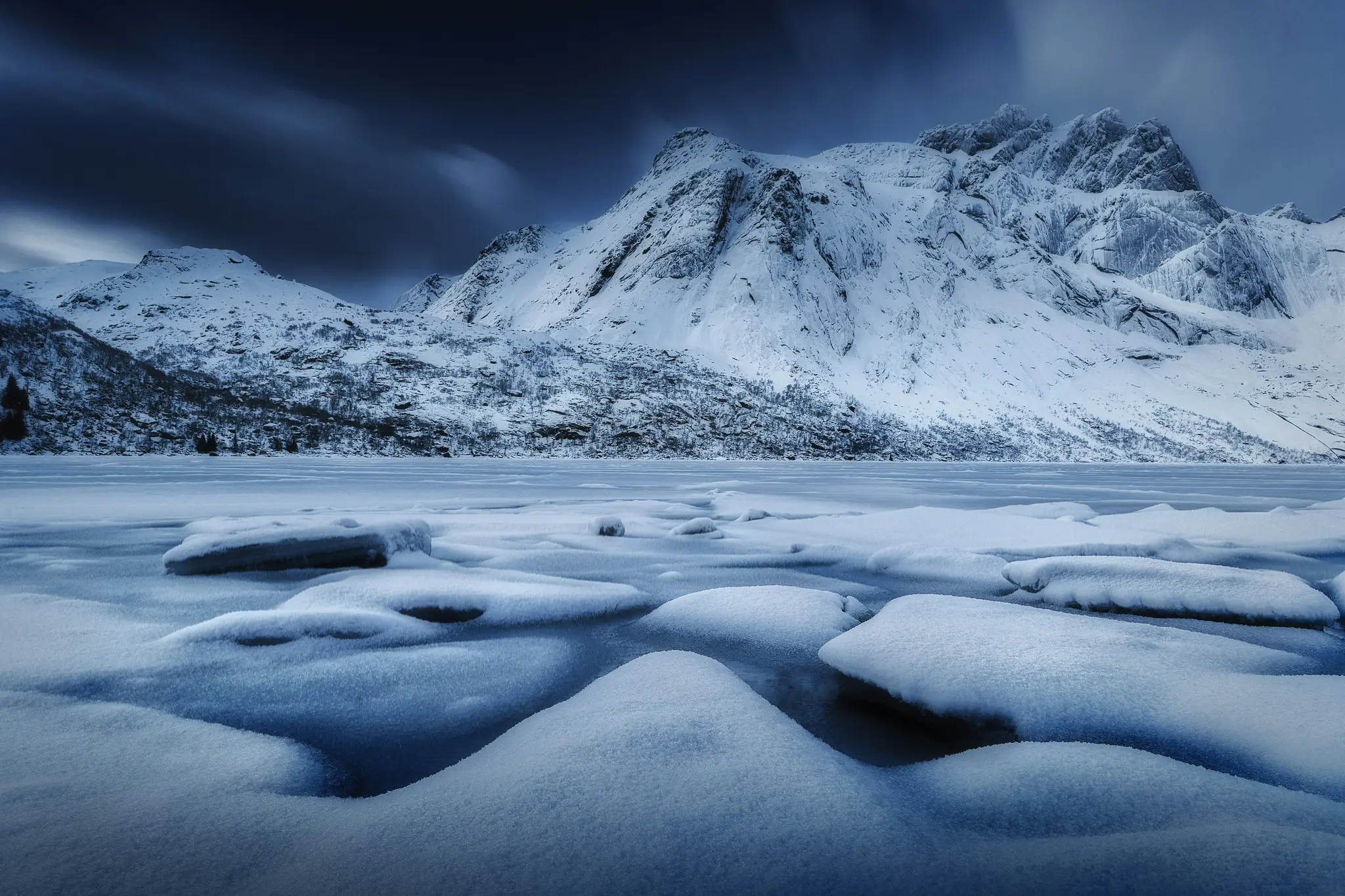 En bred landskapsvy över snöklädda berg under en mörk, molnig himmel, med en frusen sjö och snödrivor i…