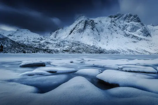 En bred landskabsudsigt over sneklædte bjerge under en mørk, skyet himmel, med en frossen sø og snedriver i…