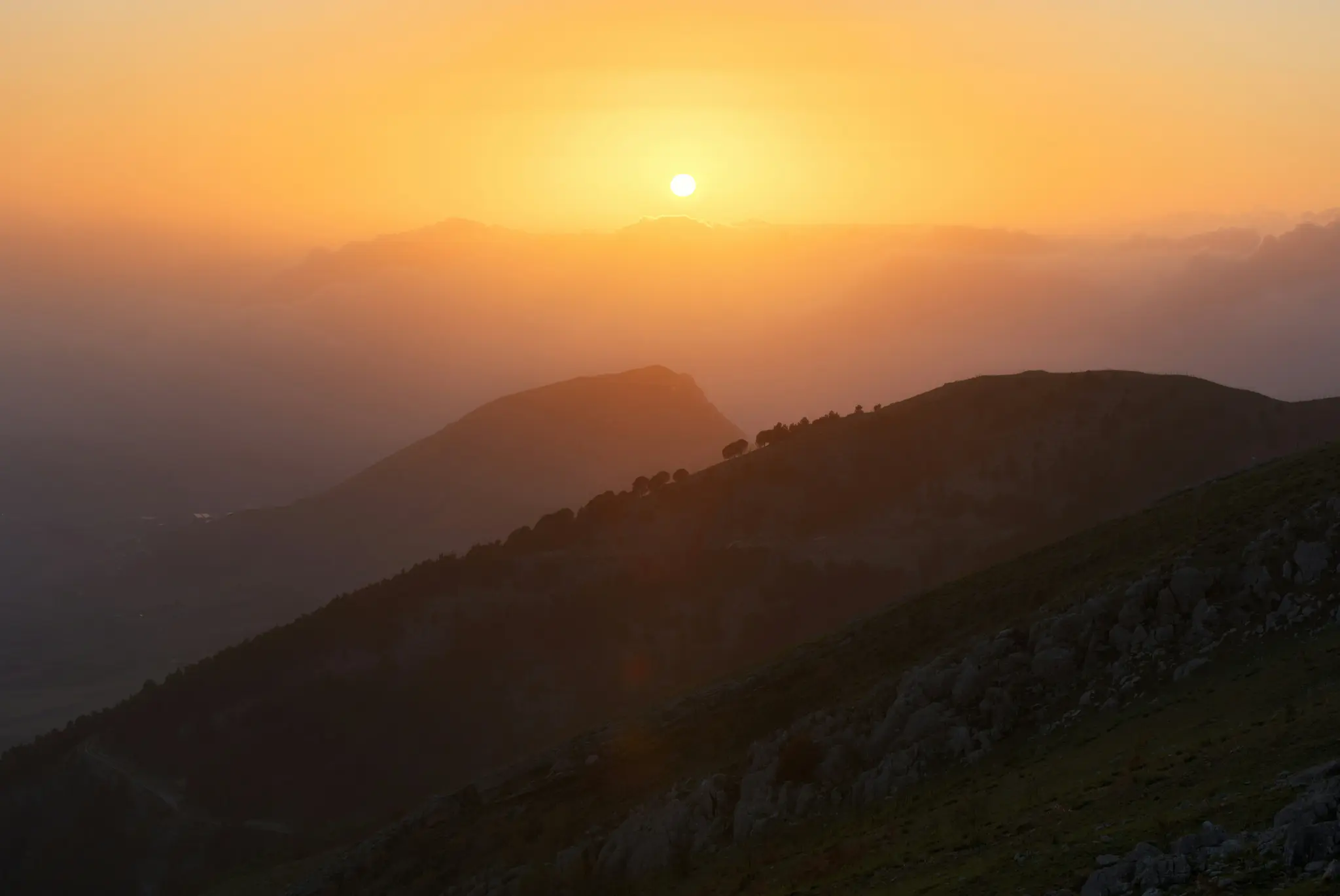 En levende orange solnedgang oplyser en bjergkæde, kaster lange skygger og silhuetterer toppene mod himlen.