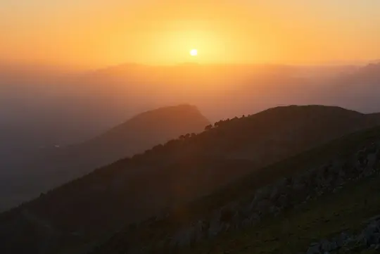 En levende orange solnedgang oplyser en bjergkæde, kaster lange skygger og silhuetterer toppene mod himlen.