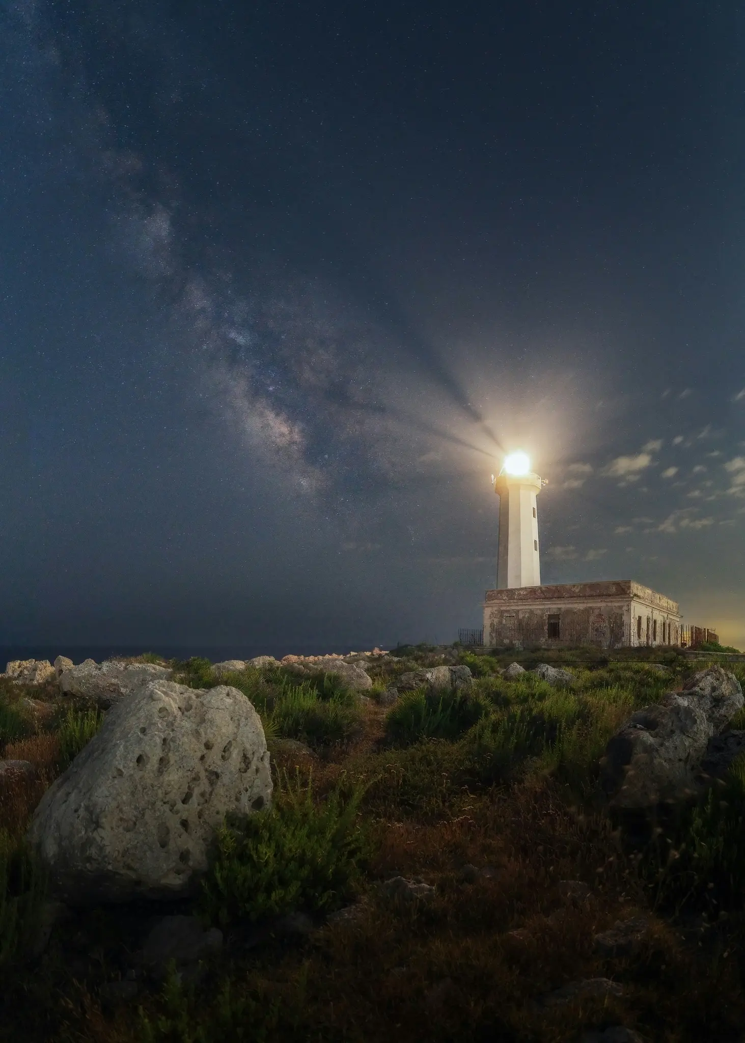 Un alto faro bianco proietta un potente fascio di luce su un cielo notturno buio e stellato dove è visibile la Via Lattea.
