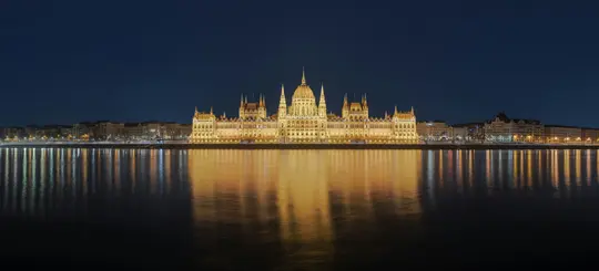 Et bredt landskabsbillede af den ungarske parlamentsbygning om natten, der er oplyst med gyldne lys og reflekteres i…