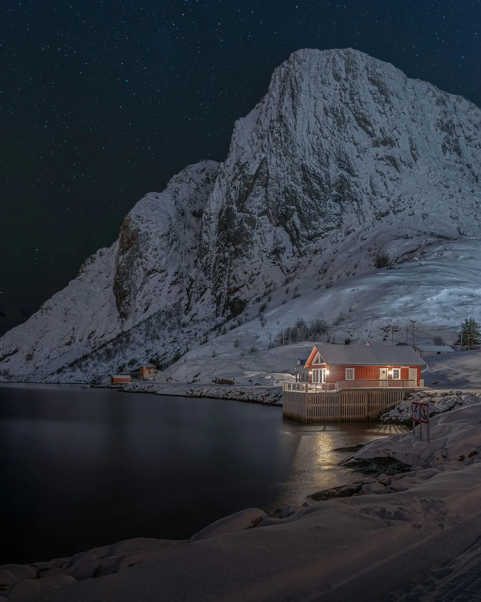Ein rotes Haus mit leuchtenden Fenstern steht am verschneiten Ufer neben dunklem Wasser und spiegelt sein Licht wider. Ein
